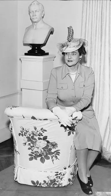 Dorothy Draper in vintage suit and feathered hat seated beside floral chair and marble bust in black-and-white interior.