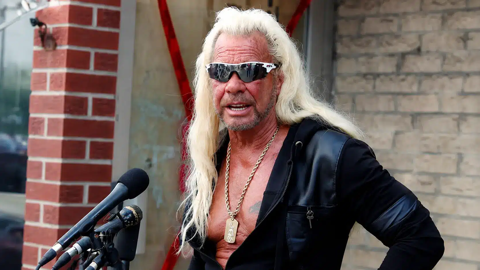 Duane “Dog” Chapman with long blonde hair and sunglasses speaking into microphones during an outdoor press event with a brick backdrop