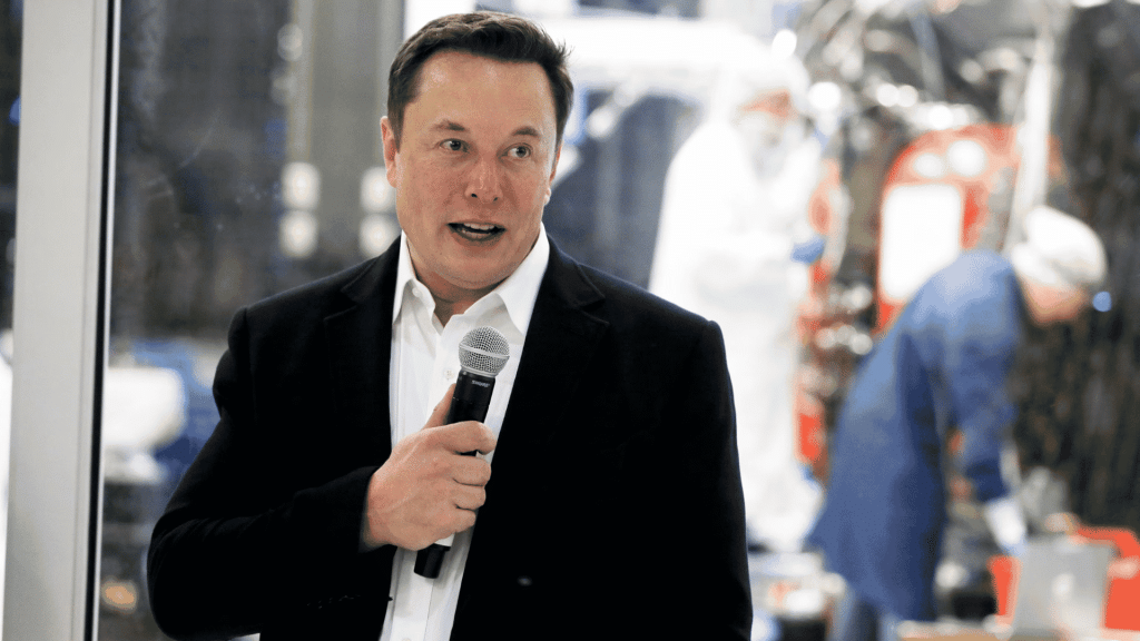 Elon Musk speaking into a handheld microphone while wearing a black blazer and white collared shirt.