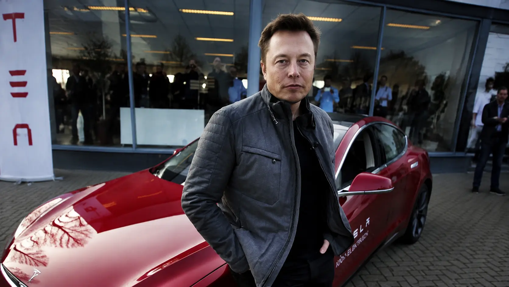 Elon Musk standing beside a red electric car outside a glass building wearing a dark jacket during a public event setting