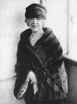 Black and white portrait of Elsie de Wolfe wearing a hat, fur wrap, and pearl necklace in a vintage style.