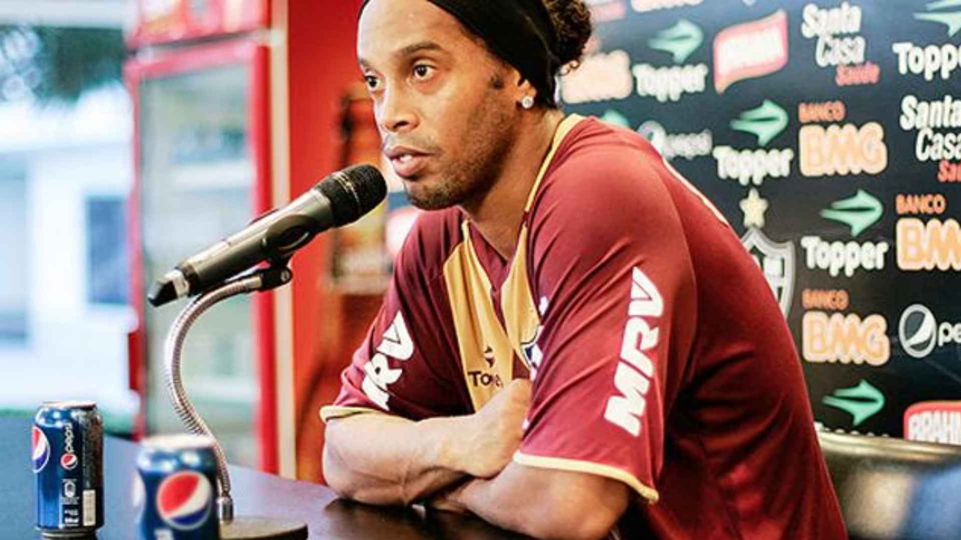 Ronaldinho speaking at a press conference wearing a football jersey with sponsor logos and a microphone in front