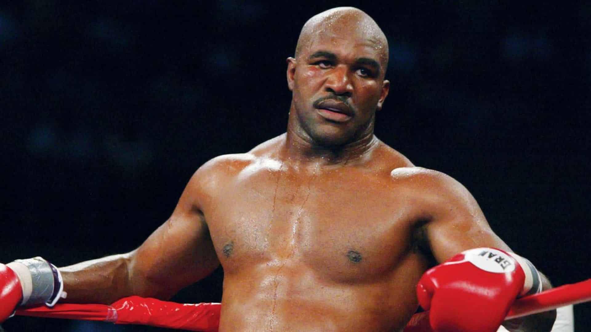 Evander Holyfield rests against the red ring ropes wearing red boxing gloves during a heavyweight fight under arena lights