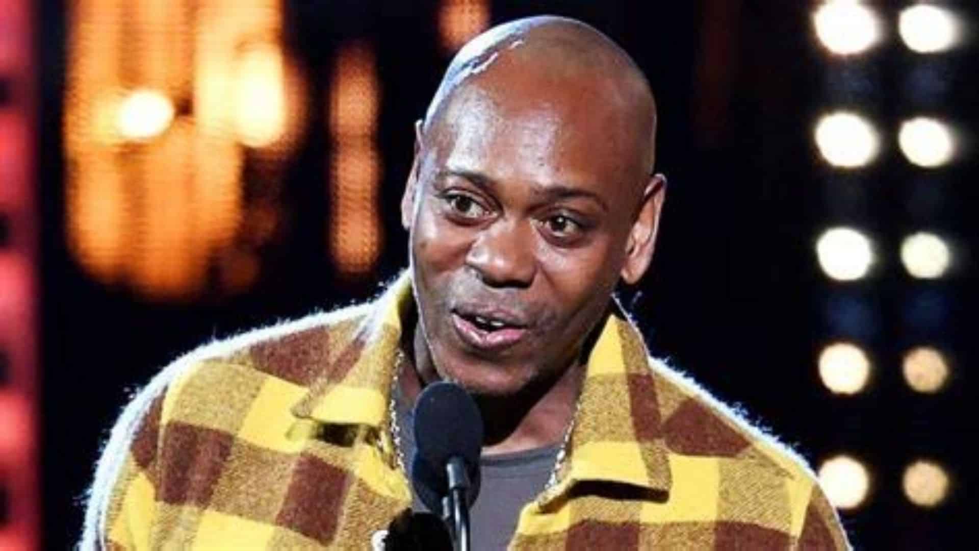 Dave Chappelle wearing a yellow and brown plaid flannel shirt jacket during a stand-up performance.