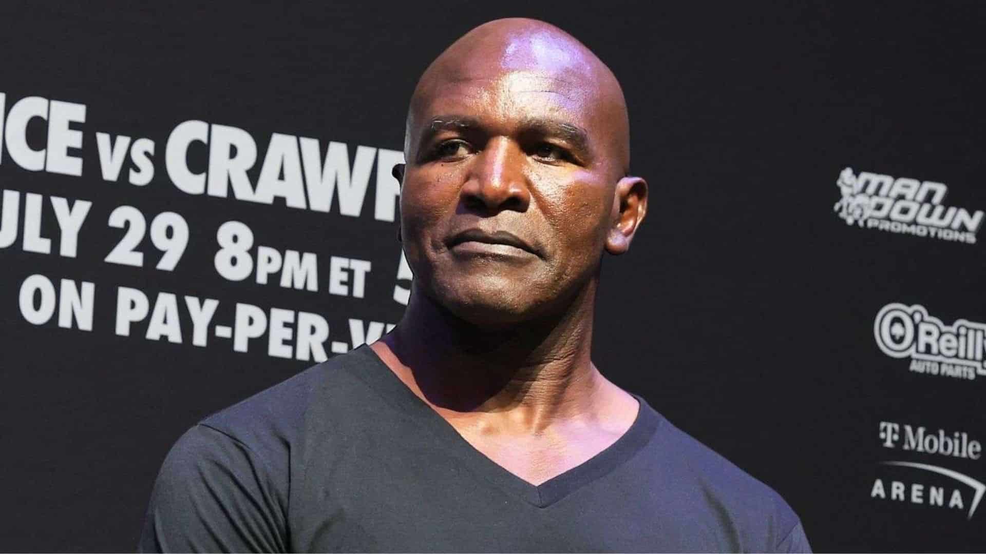 Evander Holyfield stands before a boxing event backdrop wearing a black shirt and looking to the side under bright lights