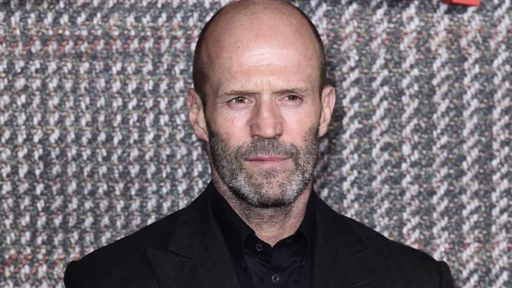 Jason Statham in black formal attire with shaved head and beard looking toward camera during a red carpet style appearance