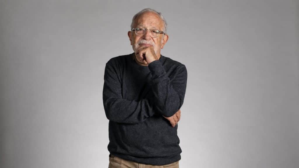 Robert Reich standing against a gray background, wearing glasses and a dark sweater.