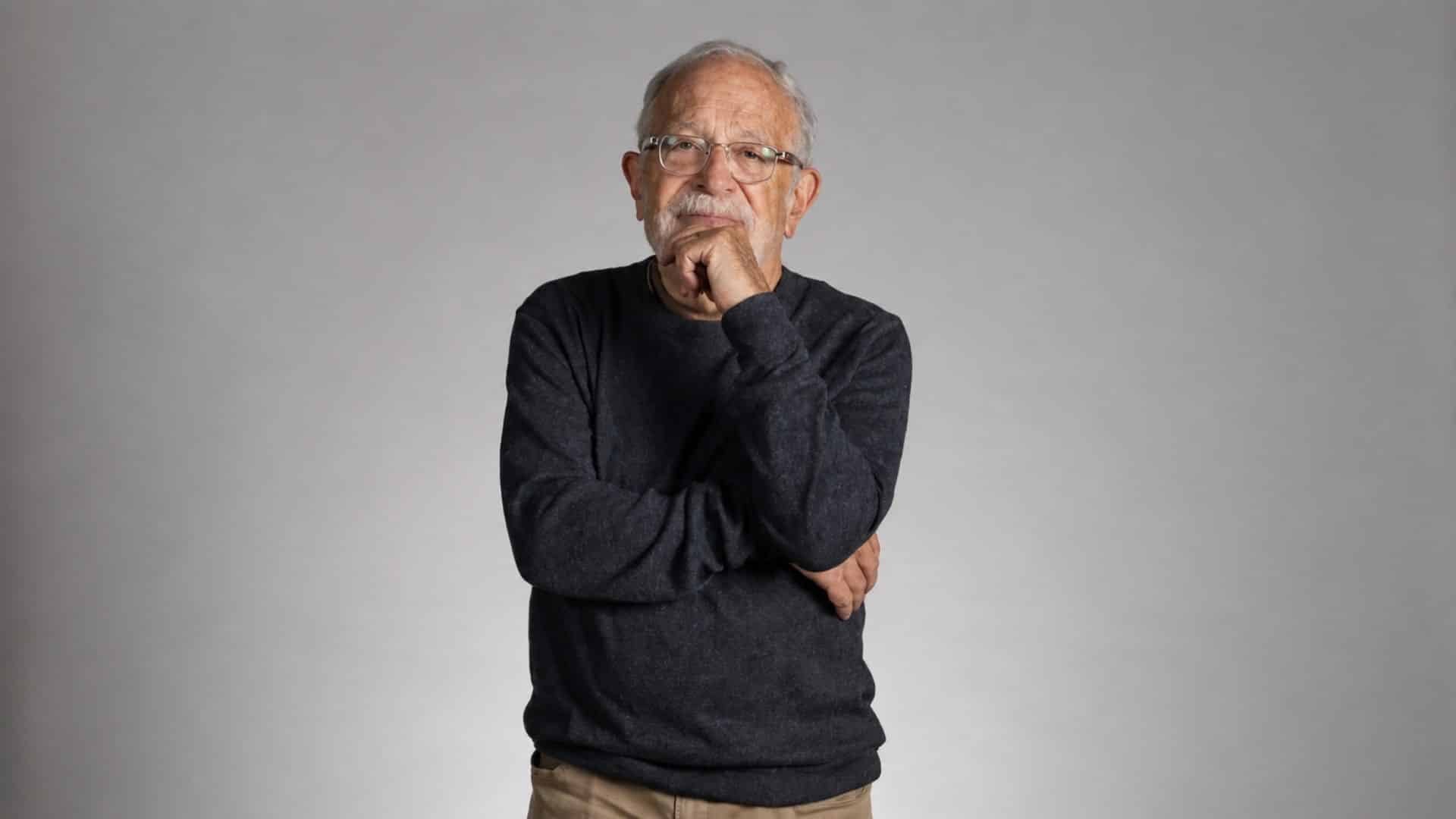 Robert Reich standing against a gray background, wearing glasses and a dark sweater.