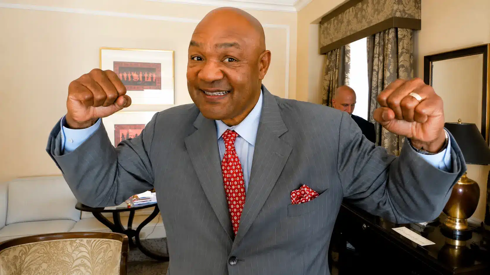 George Foreman in a gray suit and red tie raising fists indoors in a formal room with warm lighting and decor