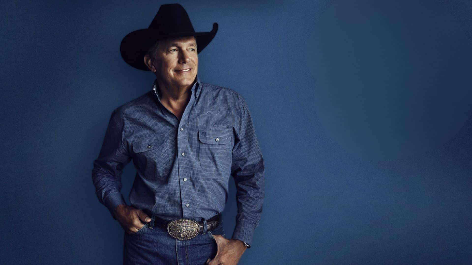 George Strait wearing a black cowboy hat and blue button-down shirt against a blue background.