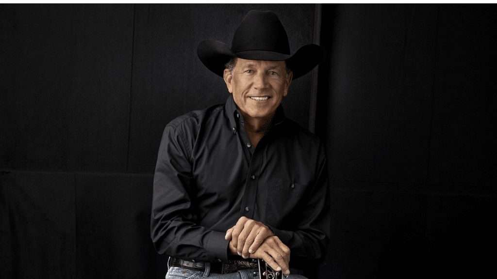 Portrait of country music singer George Strait wearing a black cowboy hat and a black button-down shirt against a dark backdrop.