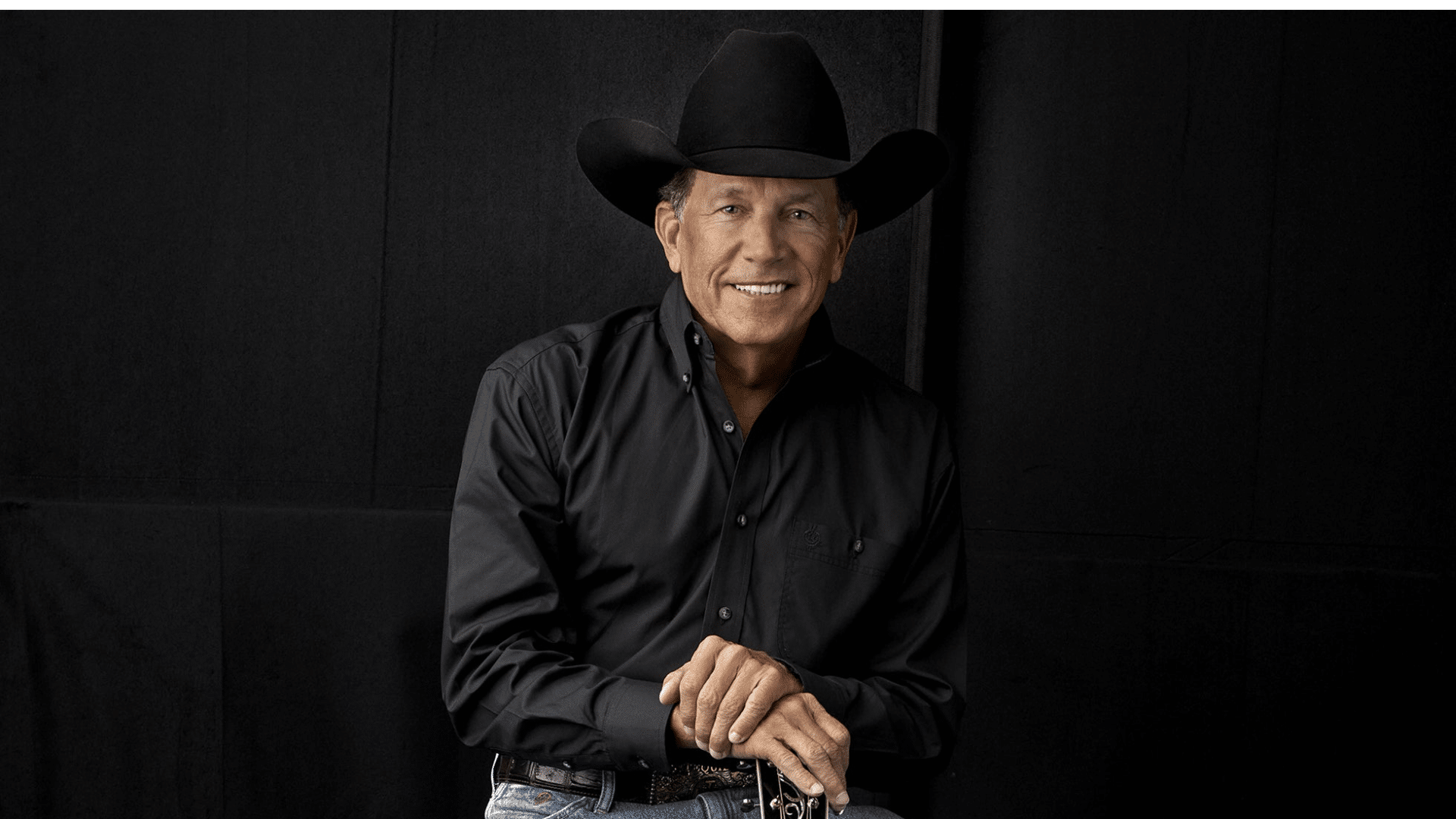 Portrait of country music singer George Strait wearing a black cowboy hat and a black button-down shirt against a dark backdrop.