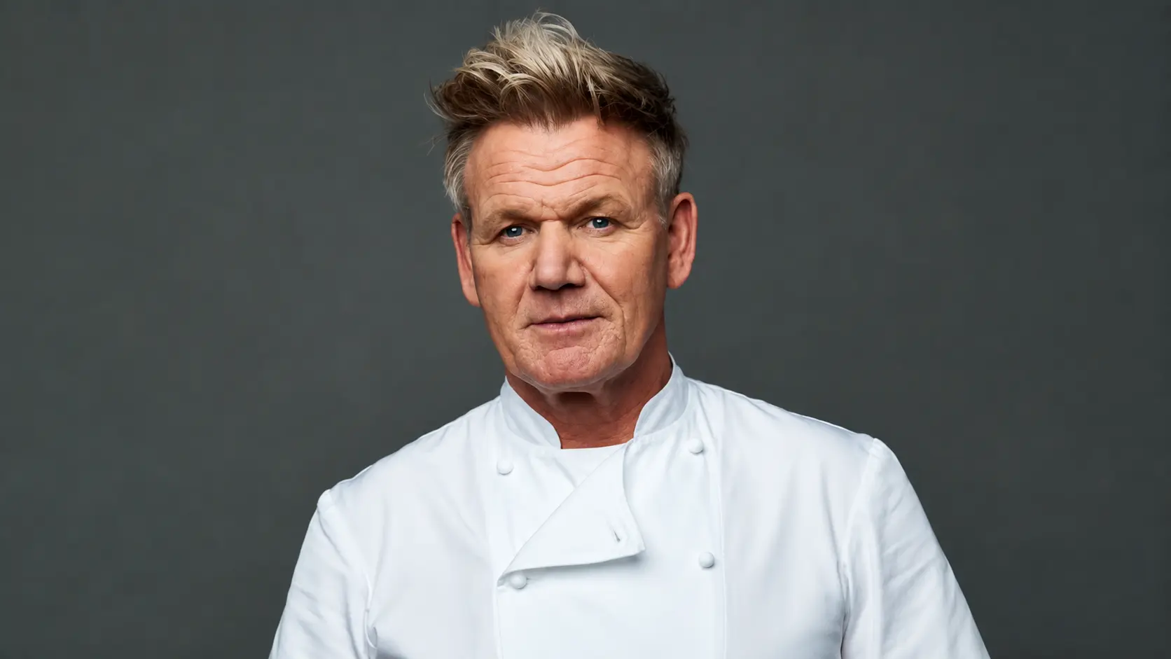 Gordon Ramsay wearing a white coat with styled hair posing for a professional studio portrait against a dark background with a calm expression
