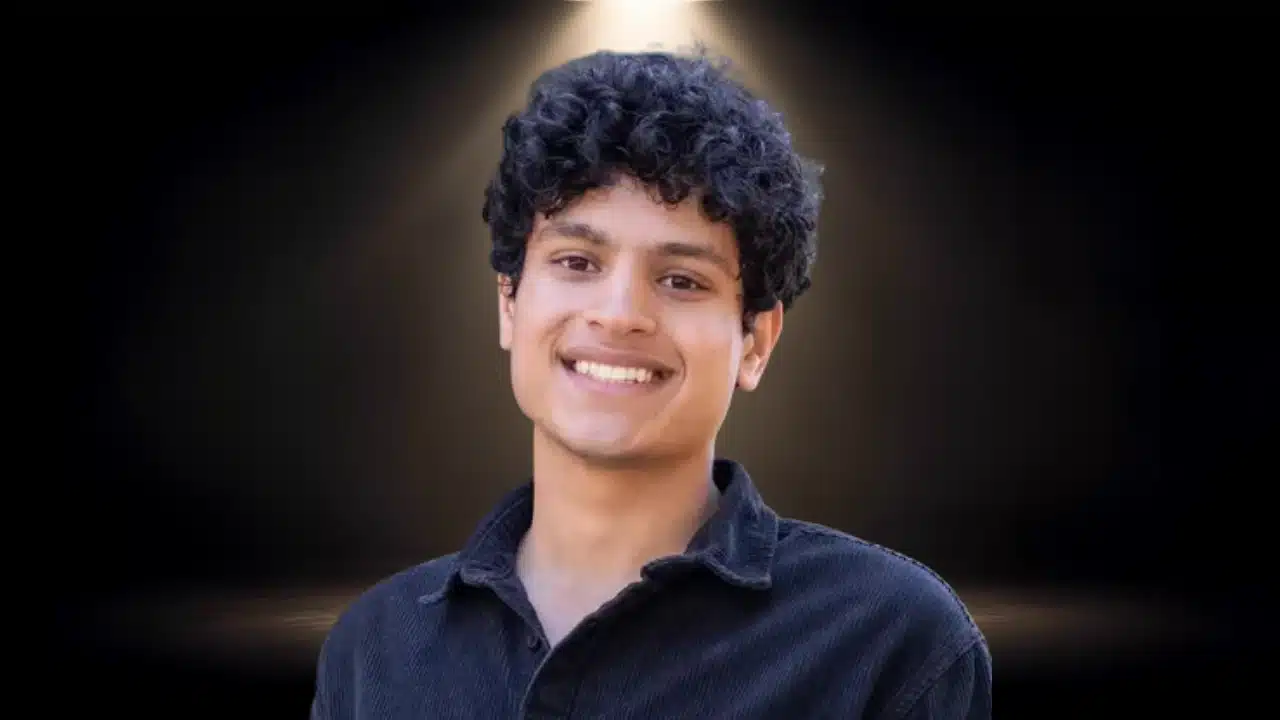 Headshot of Adarsh Hiremath smiling with curly hair, wearing a black shirt under a spotlight.