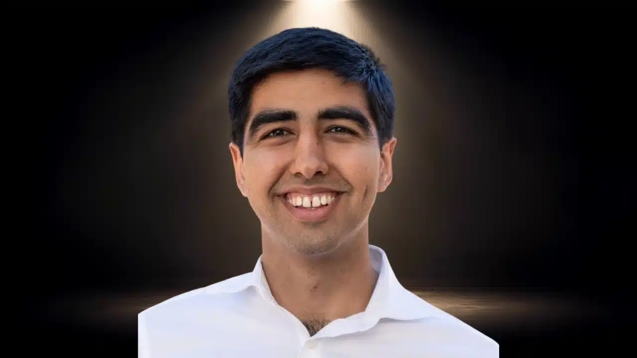 Headshot of Surya Midha smiling in a white shirt against a dark, spotlighted background.