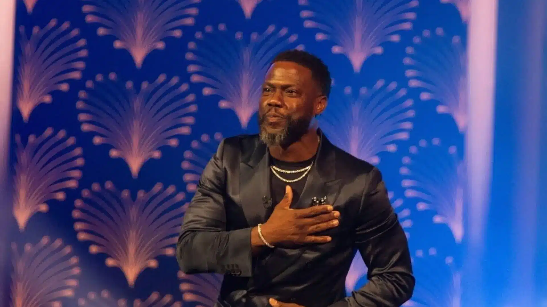 Kevin Hart wearing a black suit during a stage appearance with decorative blue lighting and a formal event setting