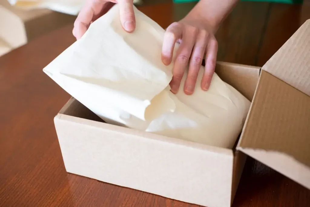 Hands packing paper-wrapped item into cardboard box on wooden table