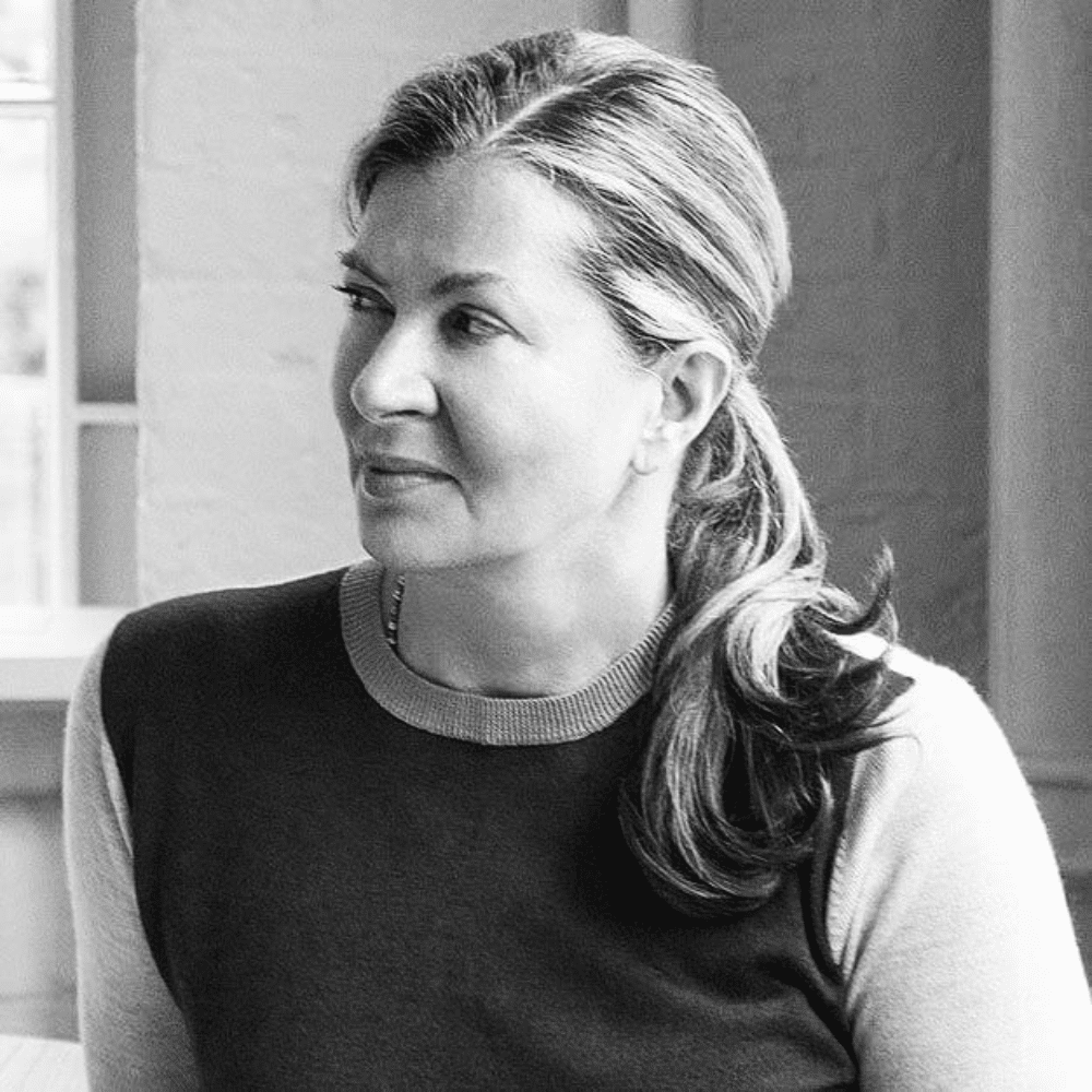 Black-and-white portrait of Ilse Crawford with ponytail wearing dark sweater, looking sideways in softly lit interior.