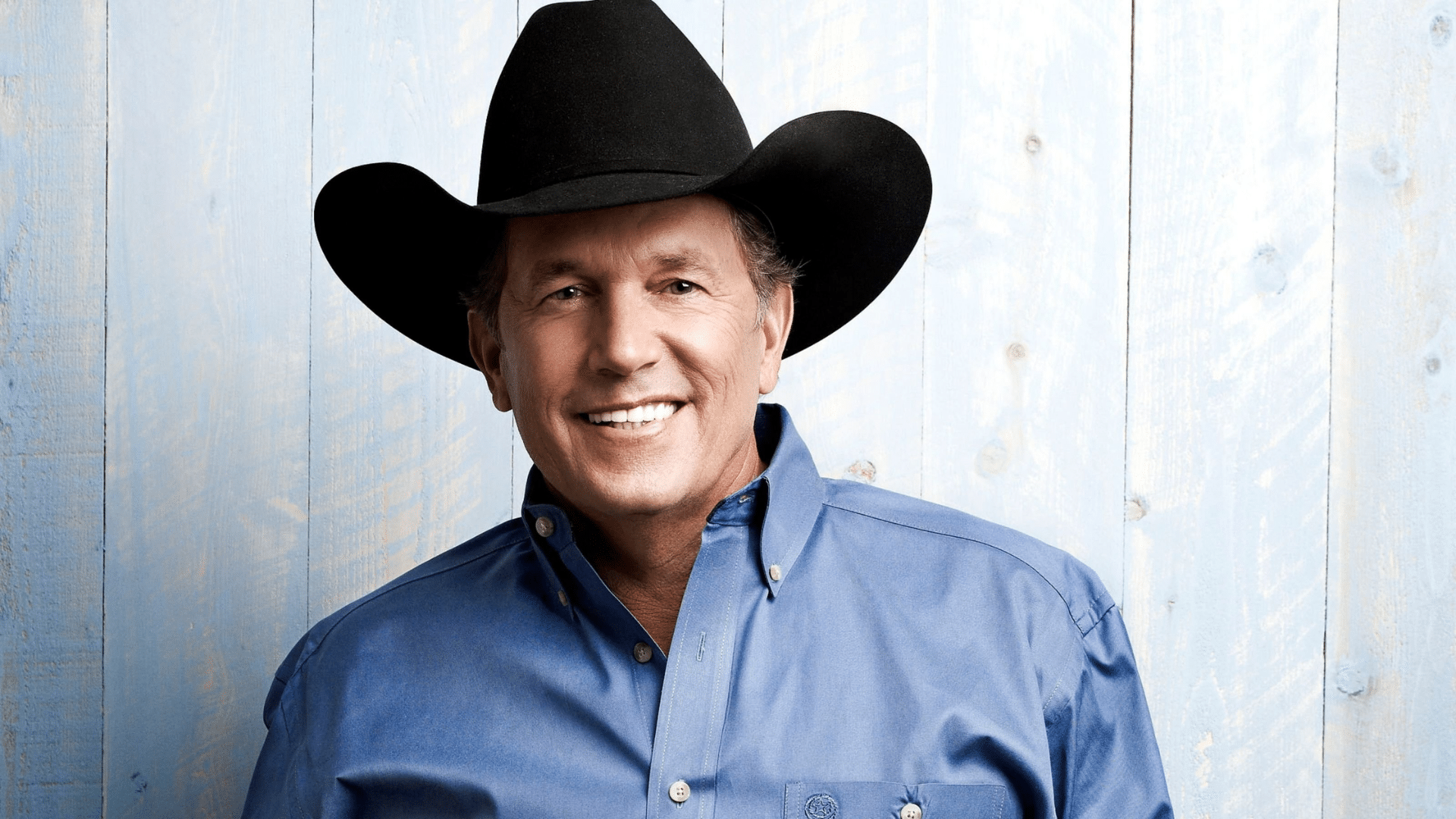 Portrait of country music singer George Strait wearing a black cowboy hat and a blue button-down shirt against a light wooden background.