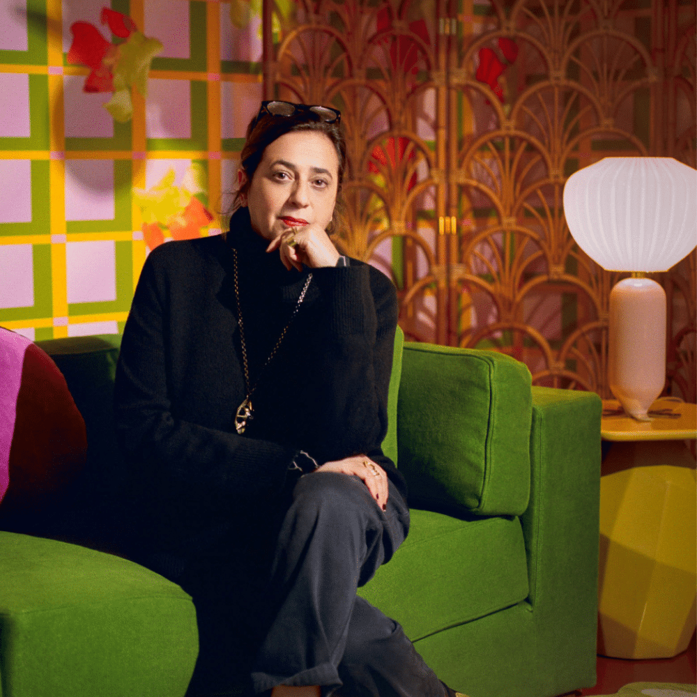 India Mahdavi seated on green sofa in colorful patterned room with decorative screen and glowing table lamp.