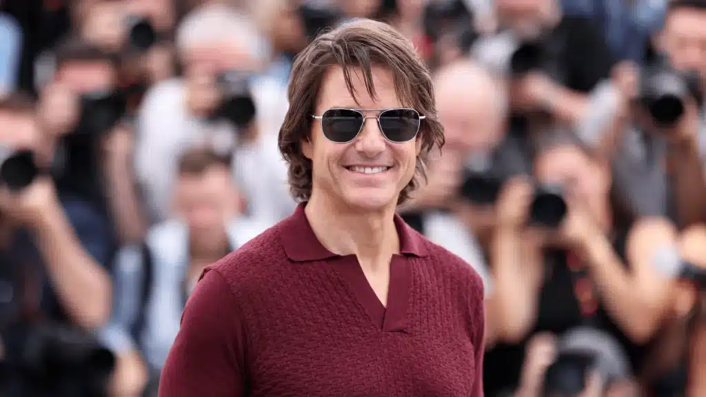 Tom Cruise smiling at a red carpet photocall, wearing sunglasses and a maroon sweater, with photographers in the background