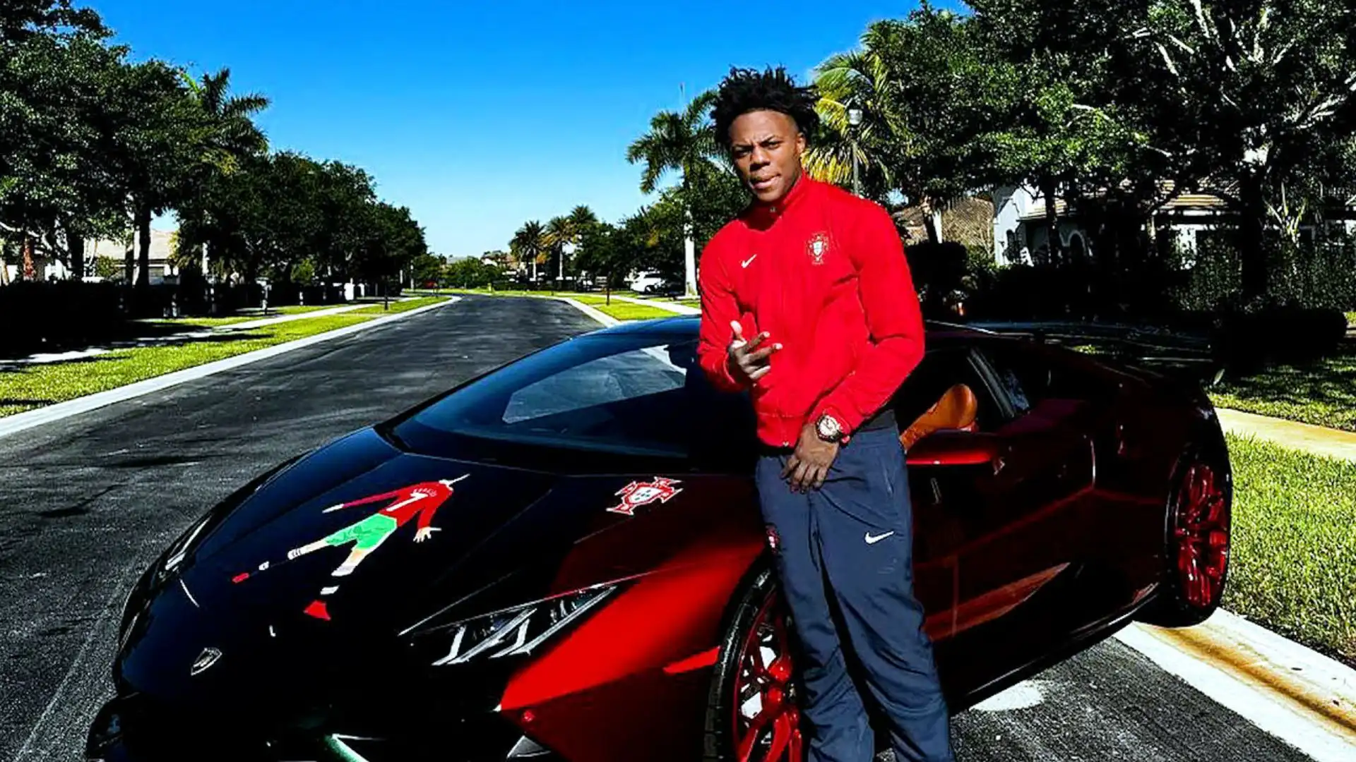 IShowSpeed posing beside a black and red Lamborghini sports car parked on a sunny residential street