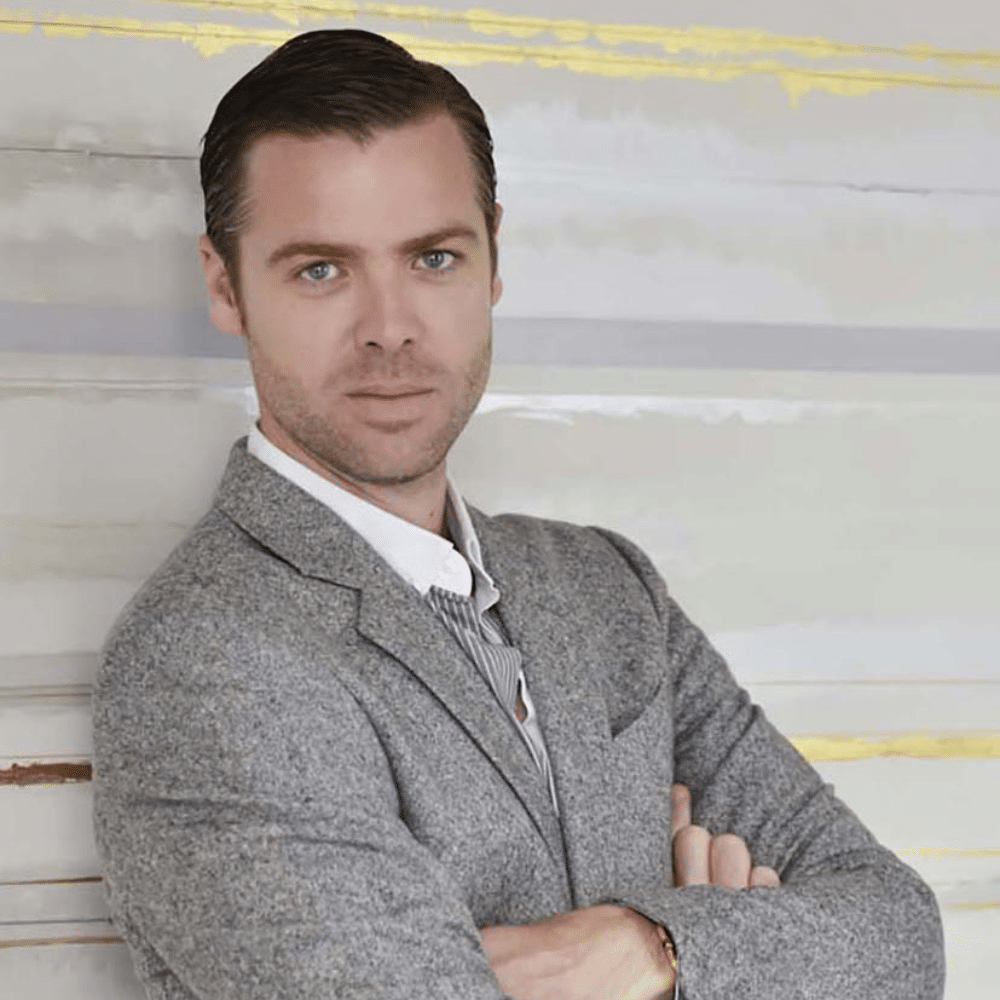 Jean Louis Denoit in gray blazer with folded arms standing against light striped wall background.