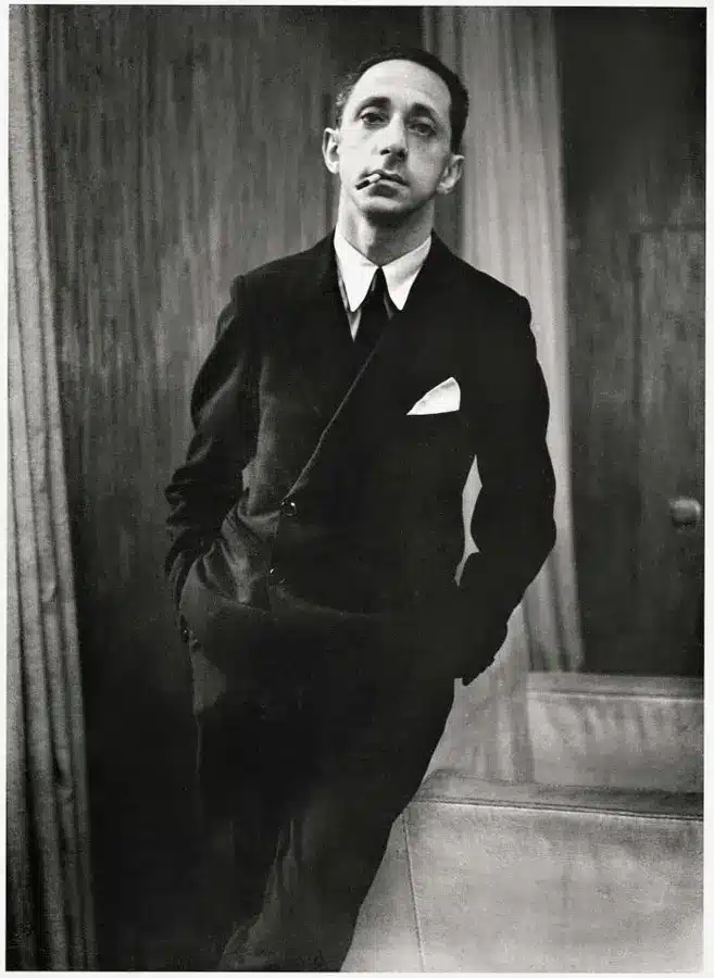 Black-and-white portrait of Jean Michel Frank in dark suit with cigarette, leaning beside sofa in formal interior.