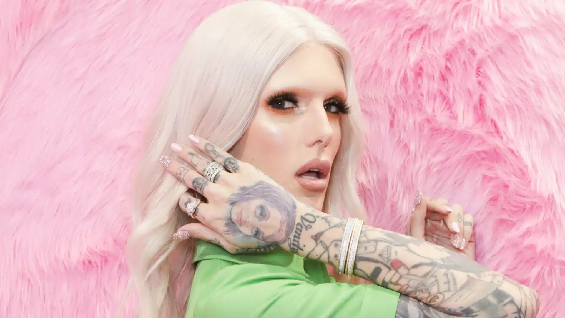 Jeffree Star with platinum hair, green top, tattoos, and makeup against pink fur