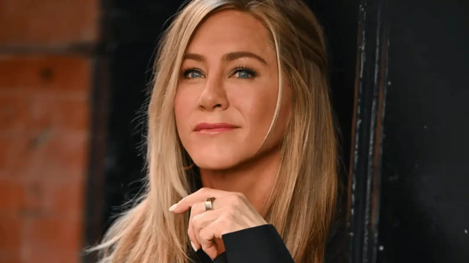 Jennifer Aniston with highlighted brown hair wearing a black outfit  with brick backdrop