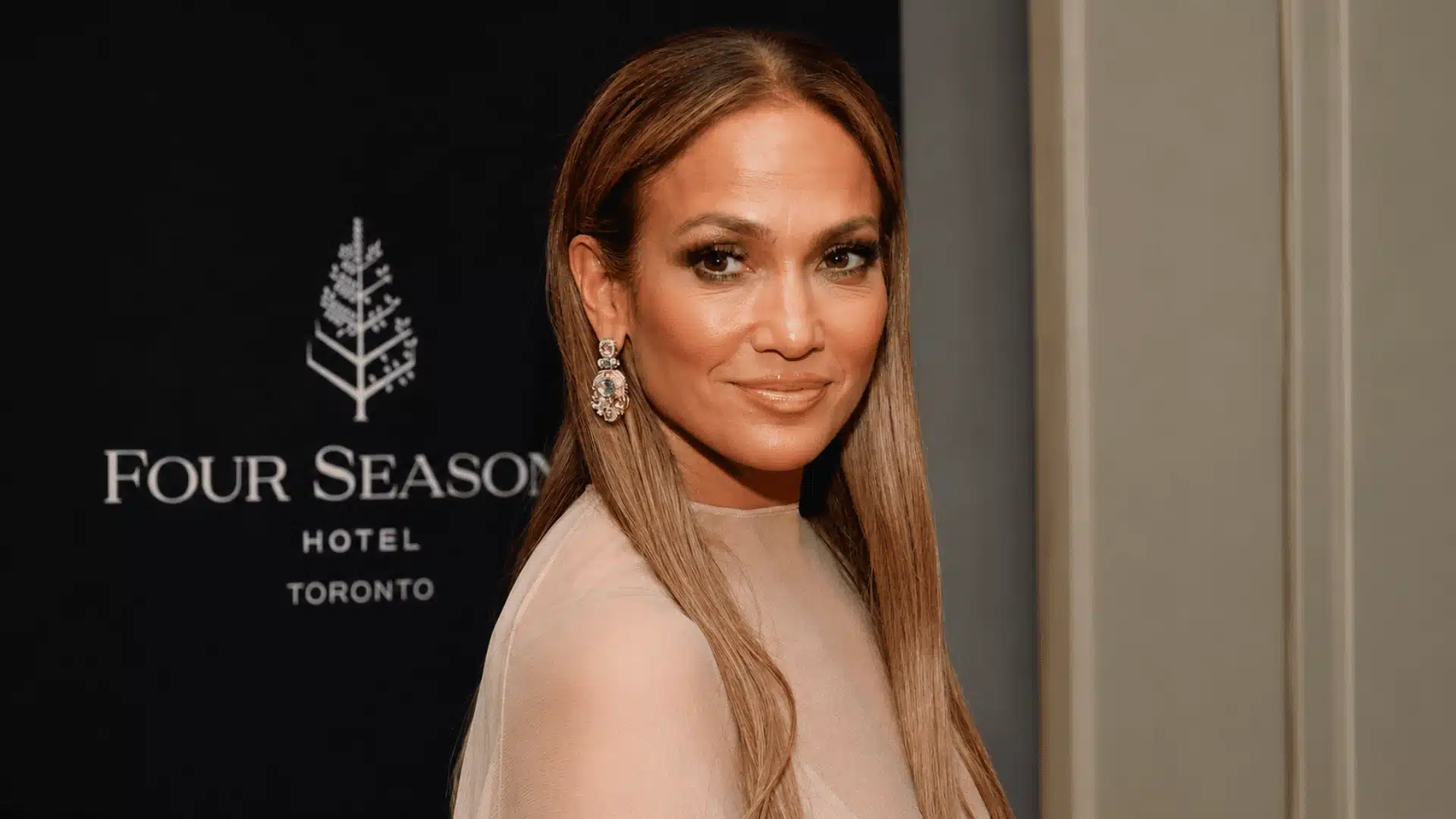 Jennifer Lopez in a beige dress with long straight hair and statement earrings posing at an event backdrop