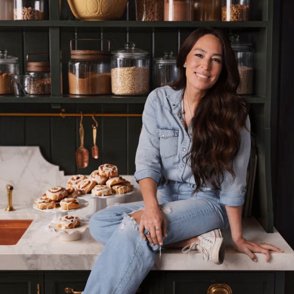 Joanna Gaines in denim outfit seated on marble countertop in modern kitchen with shelves, jars, and pastries.