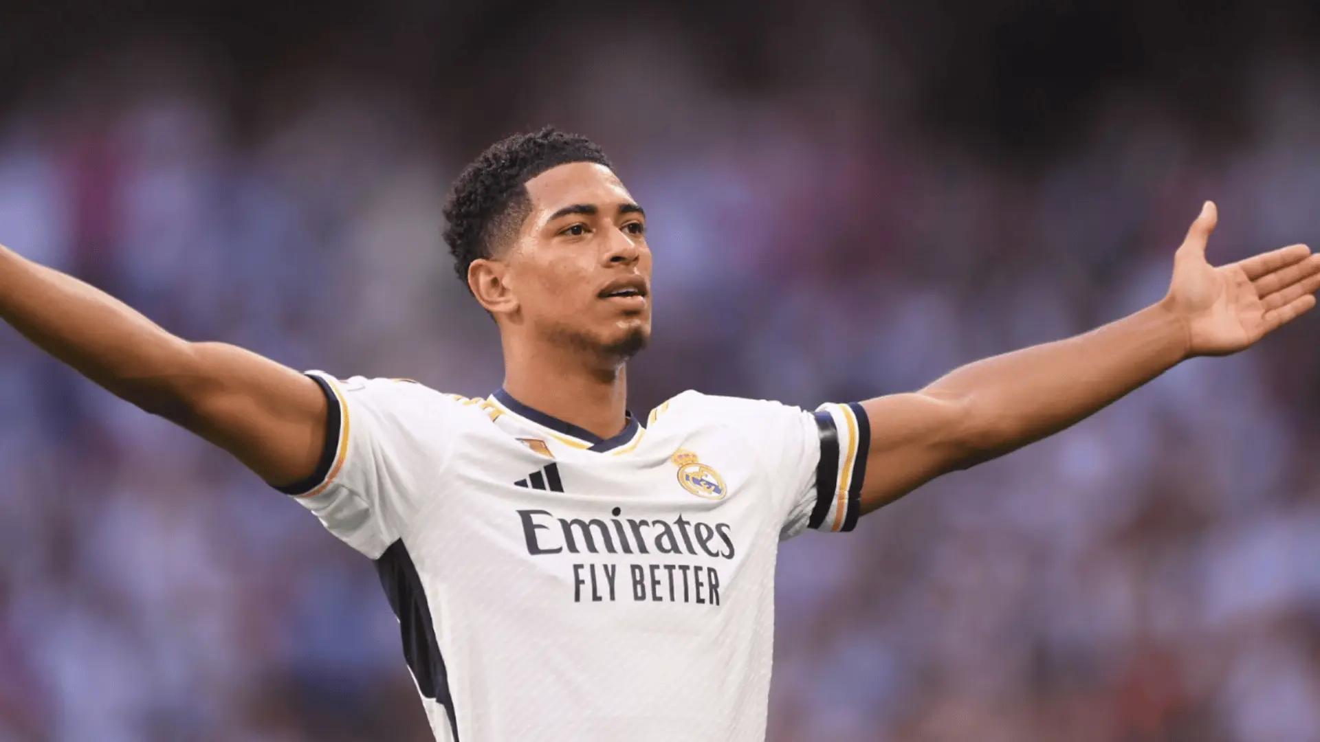 Jude Bellingham celebrates with both arms spread while wearing Real Madrid’s white soccer jersey.