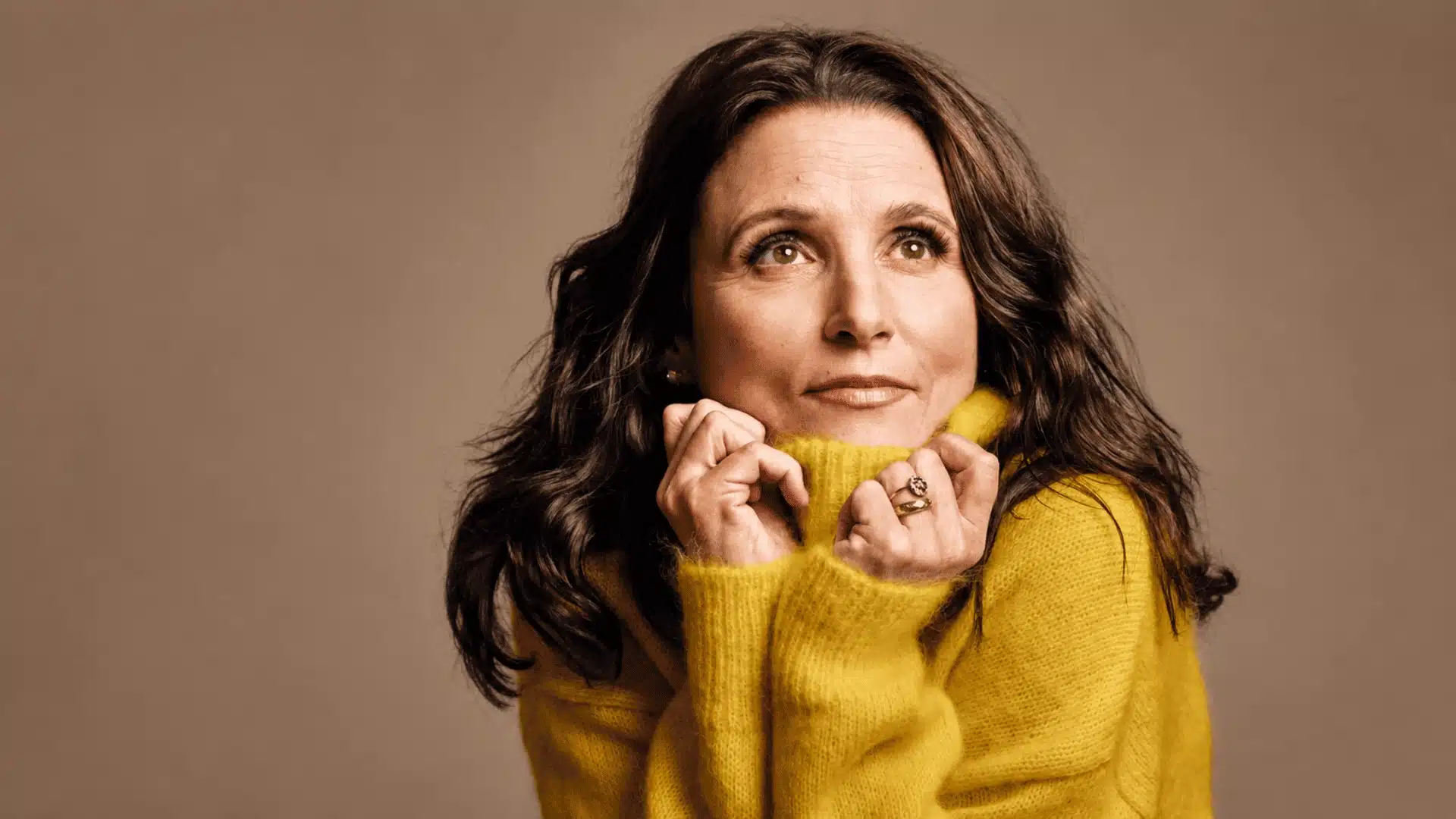 Julia Louis-Dreyfus wearing a mustard yellow sweater, posing thoughtfully against a warm beige studio background