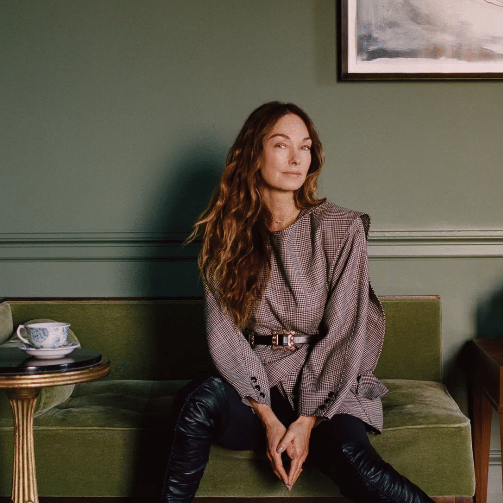 Kelly Wearstler with long hair seated on green velvet bench in stylish room with side table, teacup, and framed artwork.