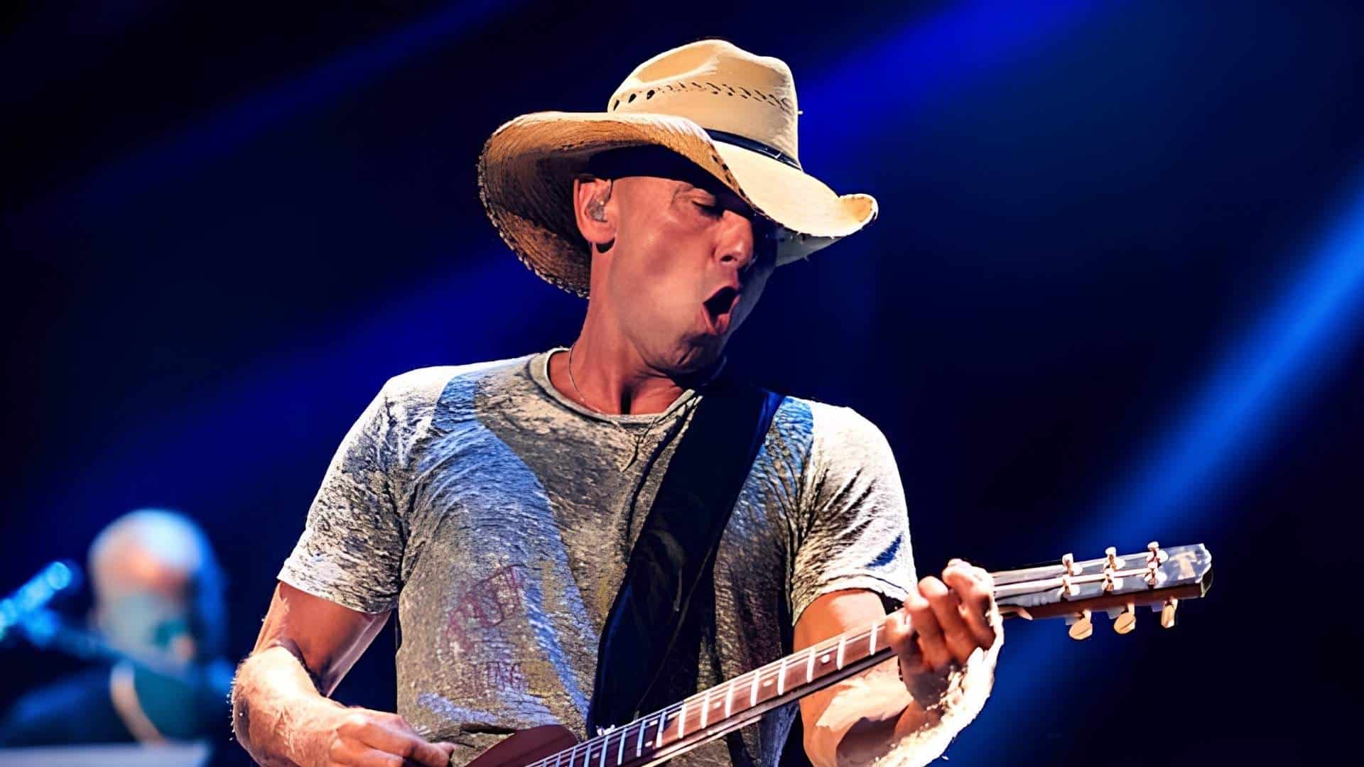 Kenny Chesney wearing a straw cowboy hat performs live on stage playing an electric guitar under blue stage lighting