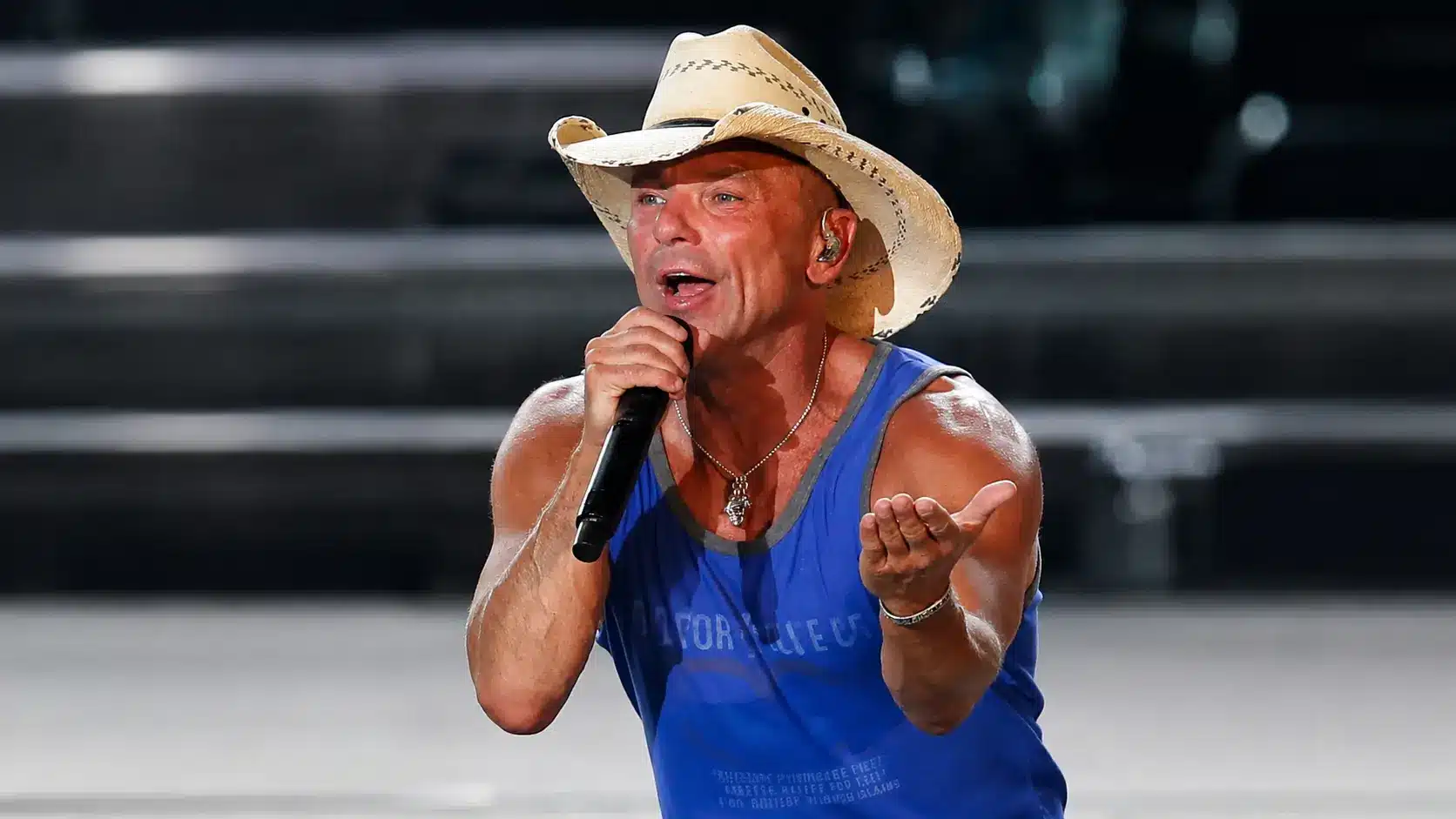 Kenny Chesney wearing a cowboy hat and blue tank top performs on stage while holding a microphone and greeting the crowd