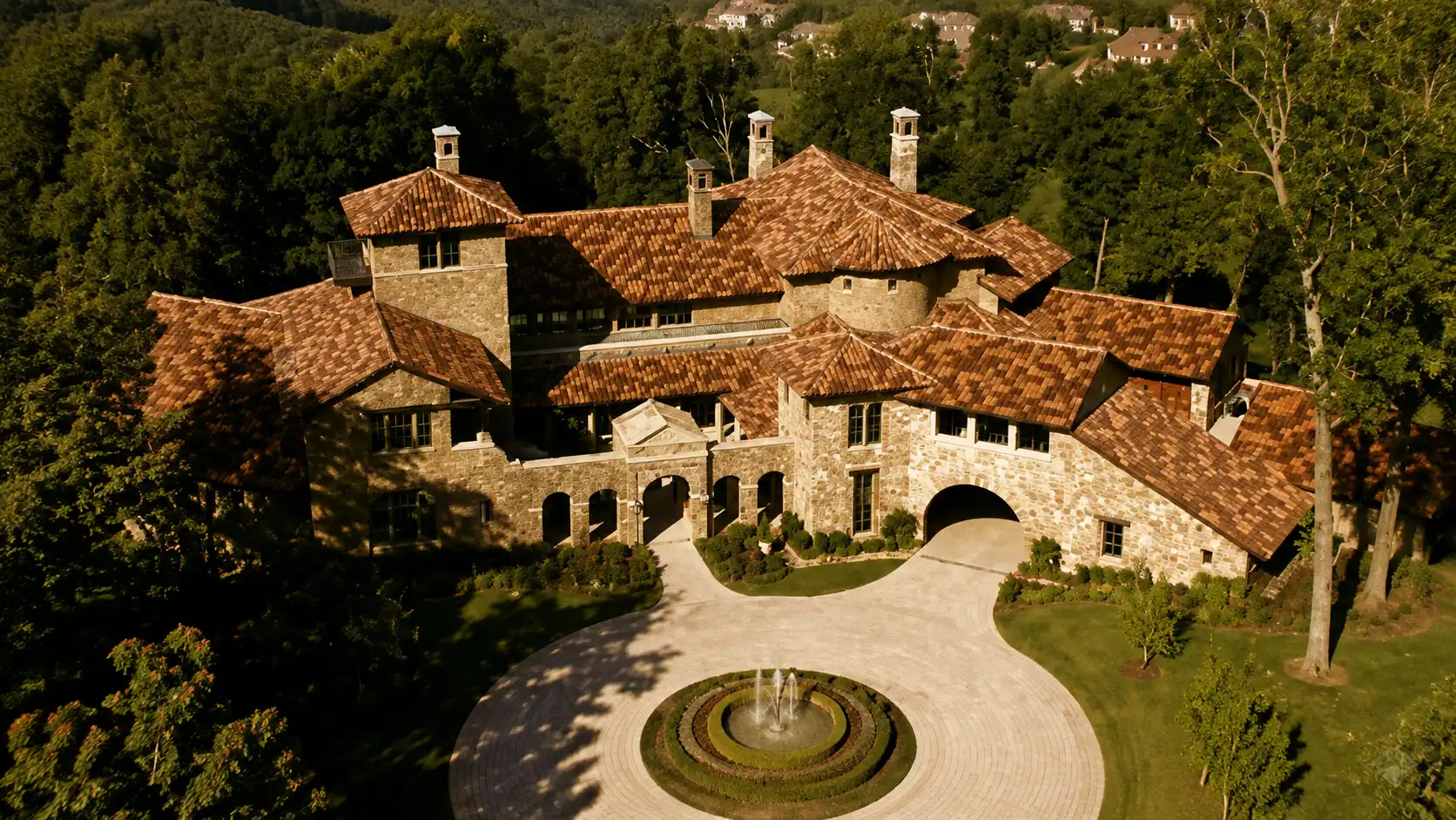 Aerial view of a large stone villa with terracotta roofs, landscaped grounds, fountain driveway, and surrounding green forest area