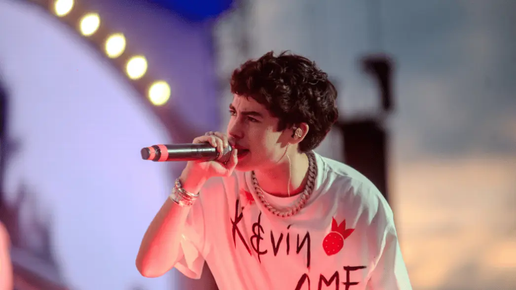 Kevin AMF performing on stage holding a microphone, wearing a white oversized graphic t-shirt and chain necklace.