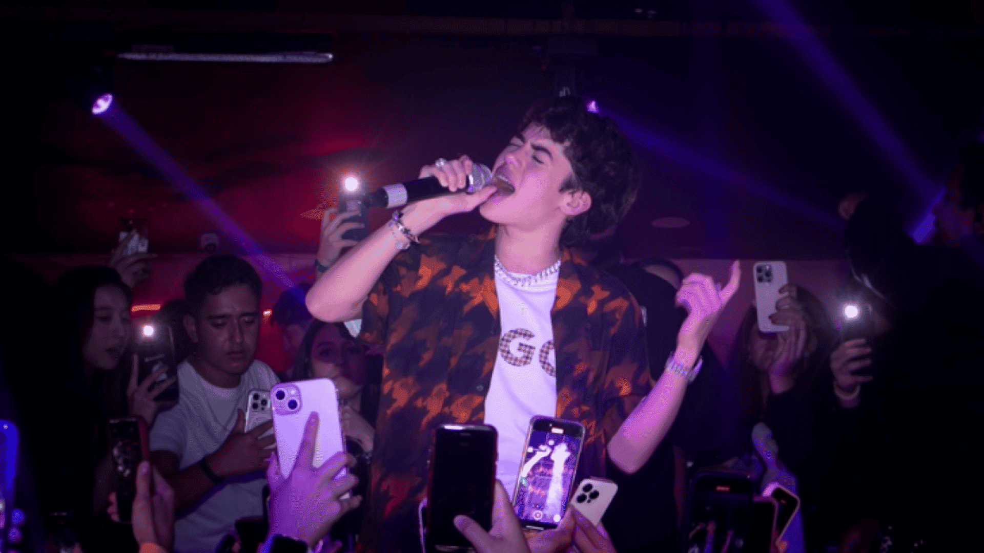 Kevin AMF singing passionately into a microphone onstage under purple lighting surrounded by fans recording on phones.