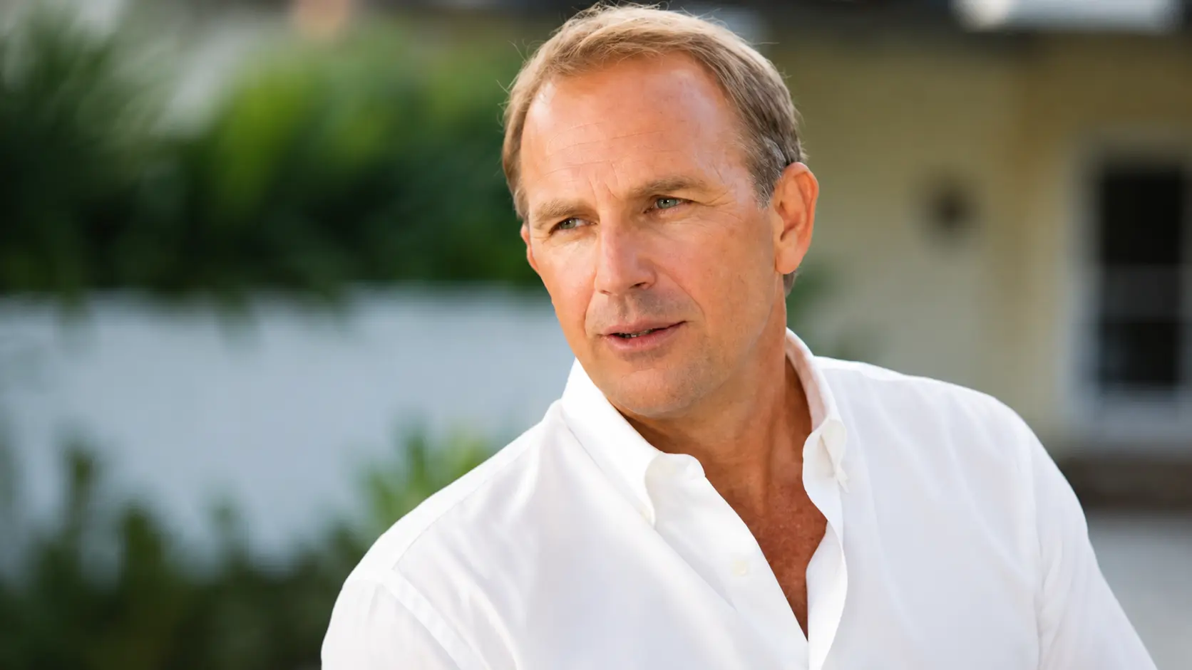 Kevin Costner in a white shirt posing outdoors with soft natural lighting and a blurred green background creating a calm portrait setting