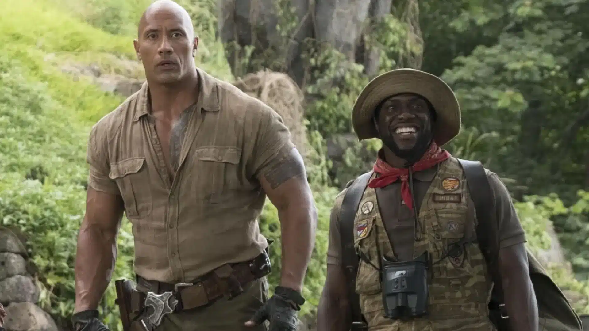 Kevin Hart appearing in an action movie scene beside Dwayne Johnson while wearing outdoor adventure clothing in a forest setting