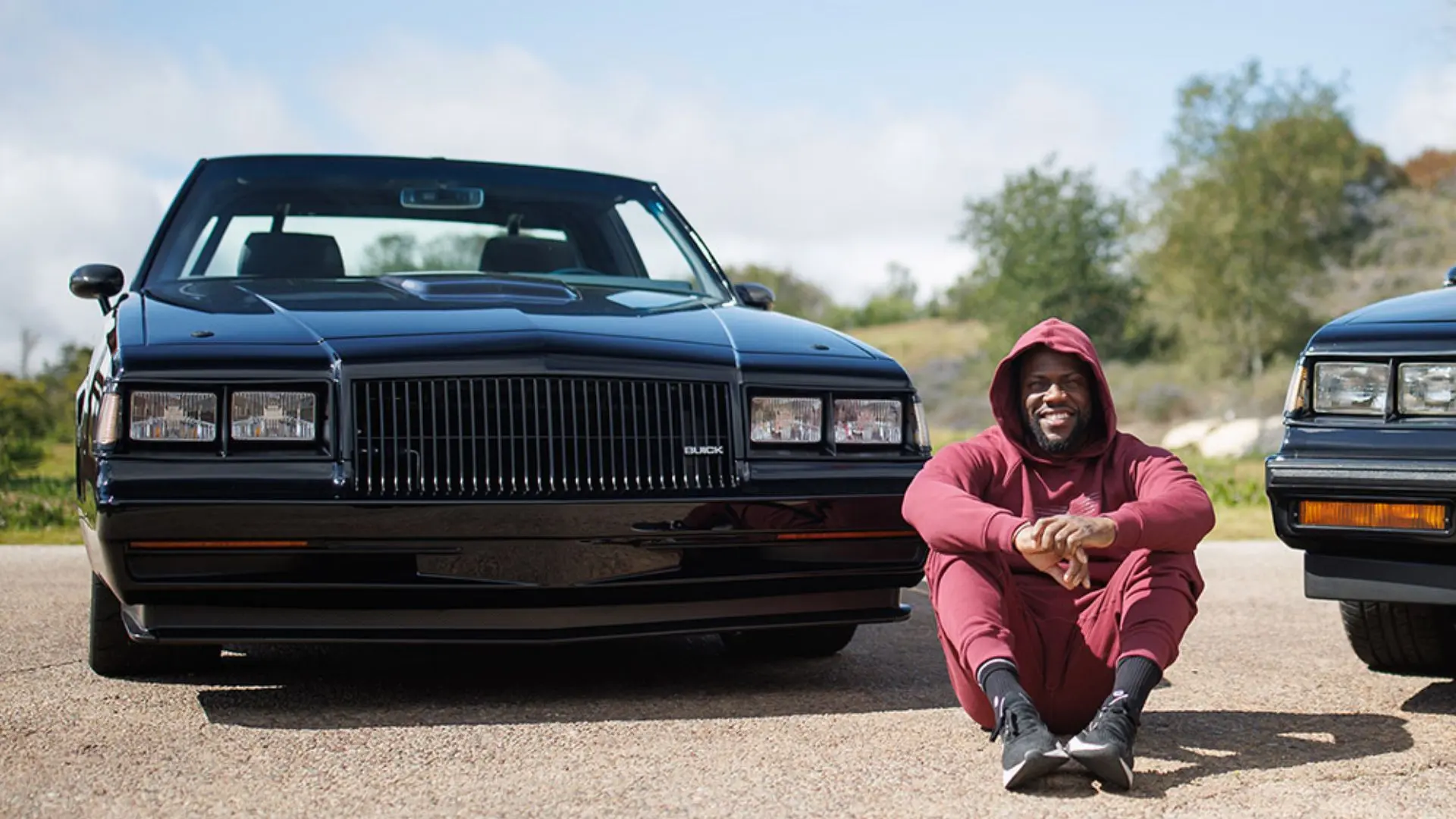 Kevin Hart sitting beside classic black cars during an outdoor automotive photoshoot with a casual maroon outfit and vintage vehicles