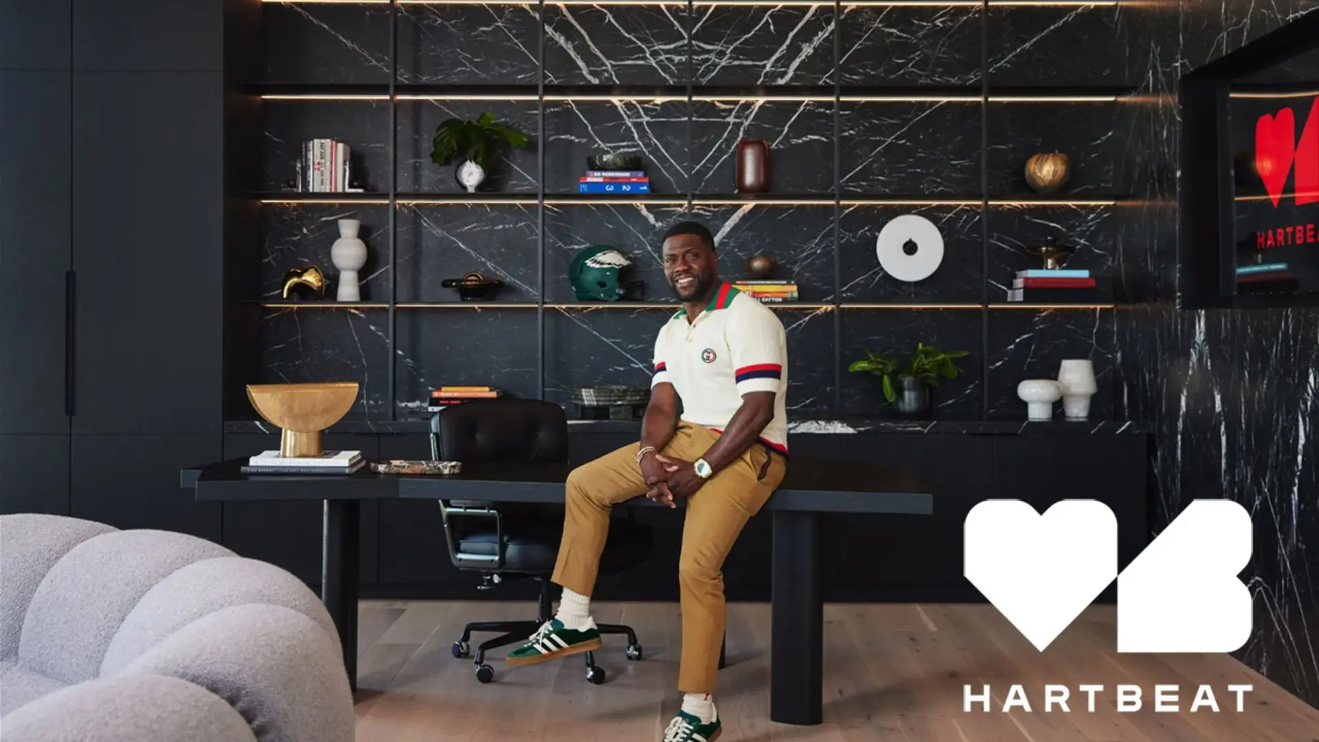 Kevin Hart sitting inside the Hartbeat office space with modern black shelves and stylish interior decorations behind him