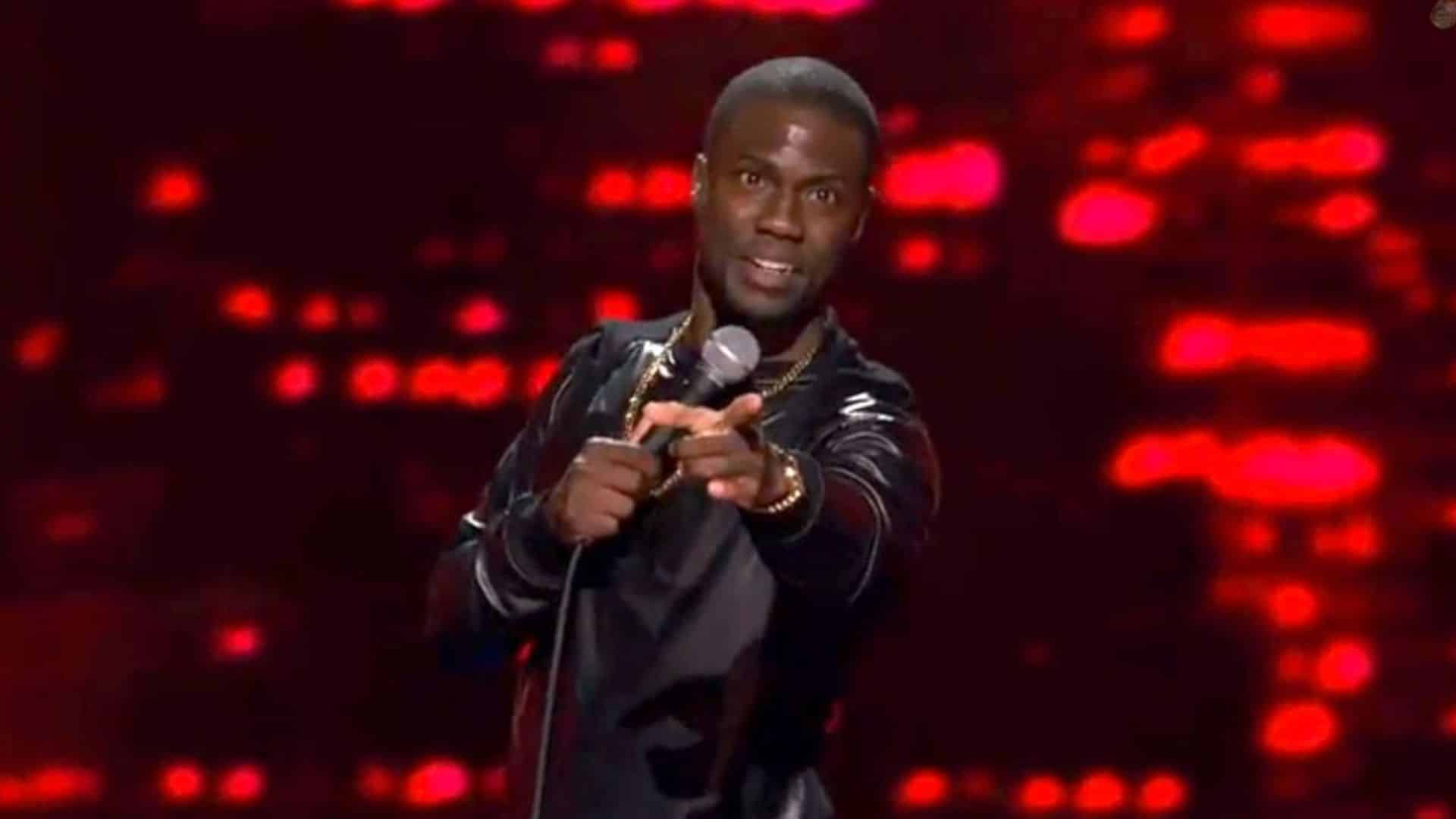 Kevin Hart performing standup comedy on stage while holding a microphone against a red illuminated background