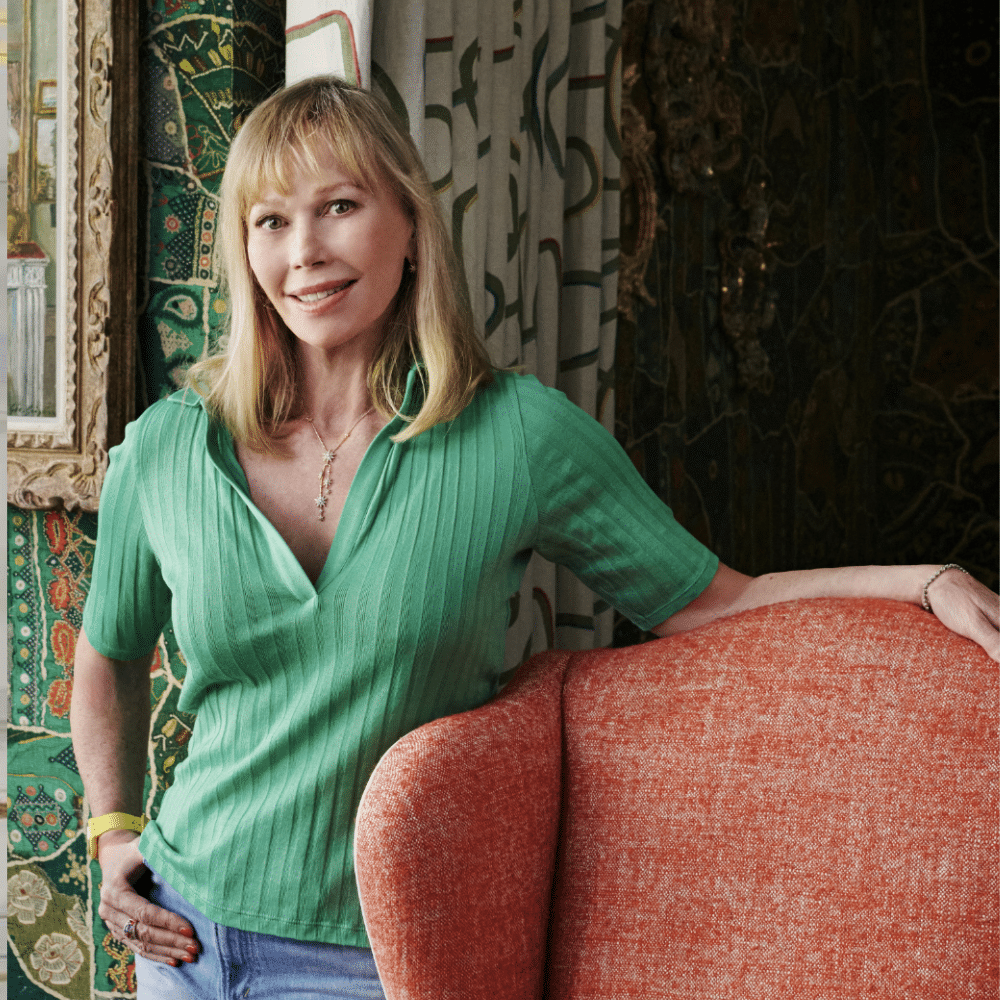 Kit Kemp in green shirt and jeans leaning on a coral chair in a colorful, patterned living space.