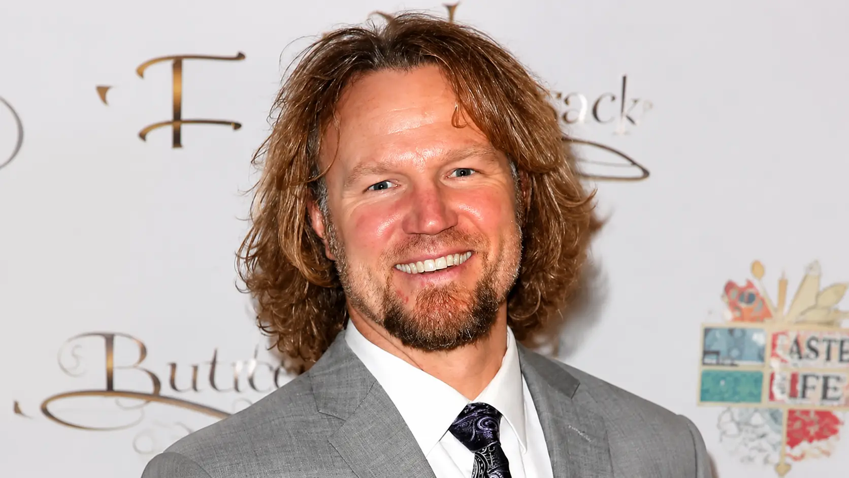 Kody Brown wearing a gray suit and patterned tie at an event with a white backdrop and shoulder length hair setting