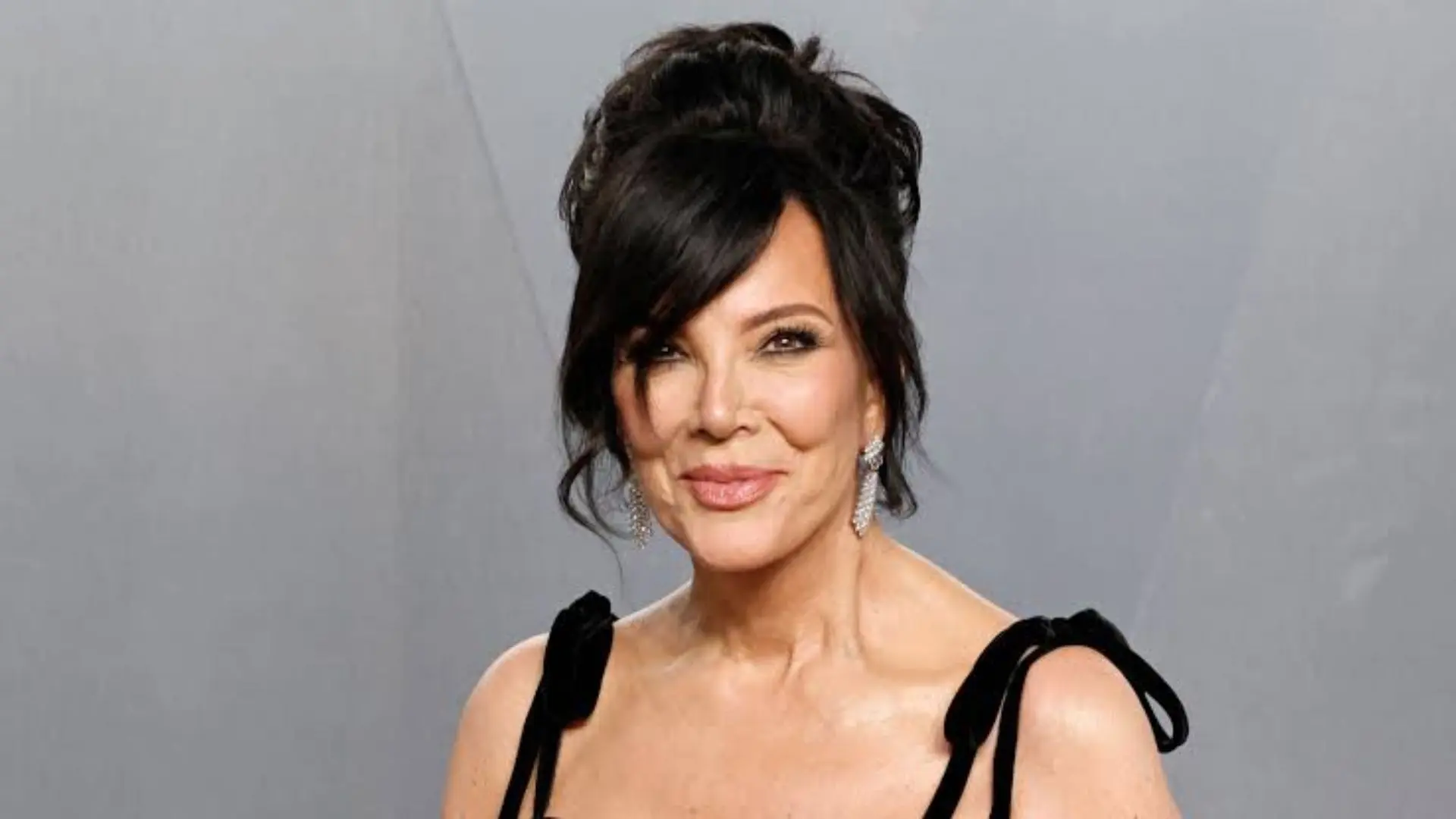 Kris Jenner wearing a black outfit with styled hair and earrings posing against a plain gray background at an event