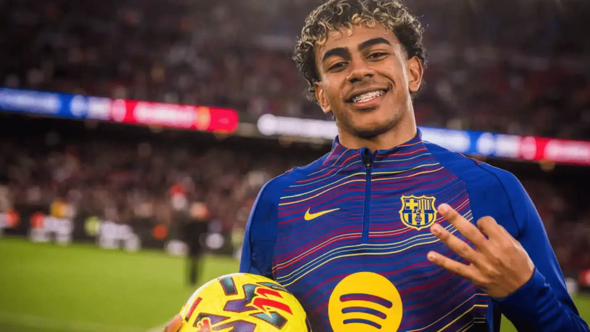 Lamine Yamal smiles while holding a match ball and wearing a Barcelona training jacket on the soccer field.