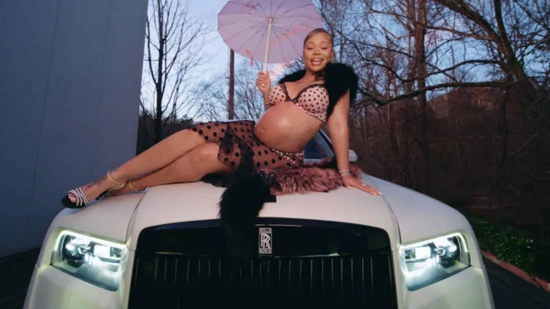Latto posing on a white luxury car during a music video shoot while holding a pink umbrella.